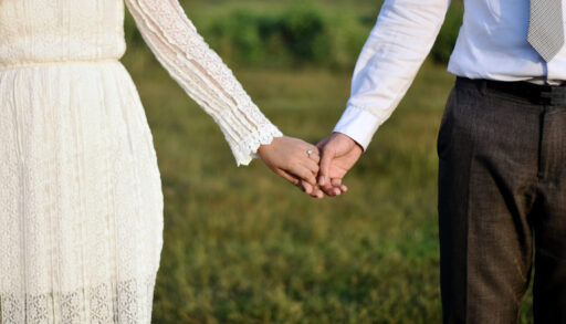 Formal attire romantic couple holding hands together while wearing engagement or wedding rings with their hands in a beautiful nature soft blur background illuminated by the gorgeous sun.