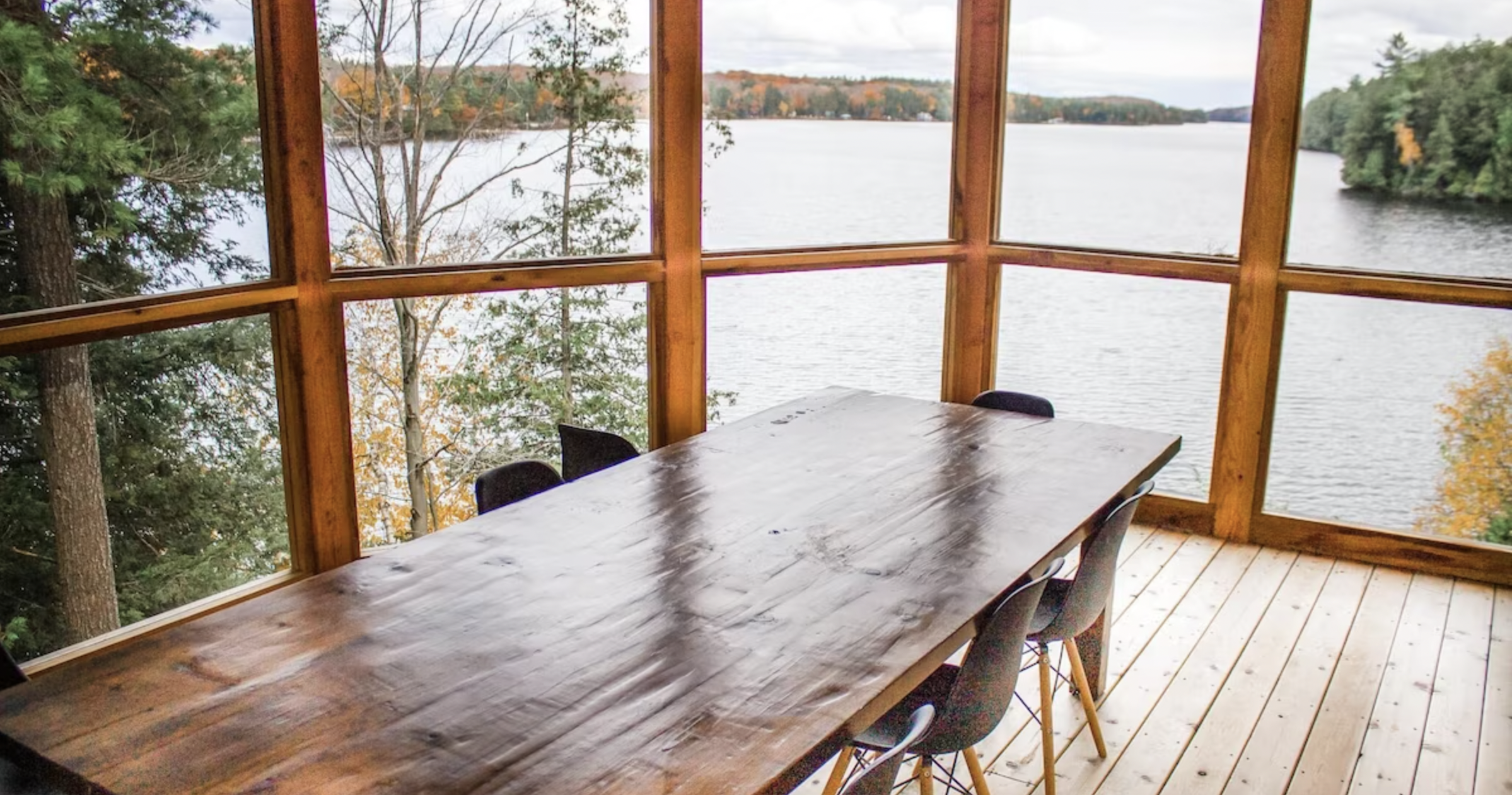 a wood table with chairs around it in a room with window walls looking out over the lake