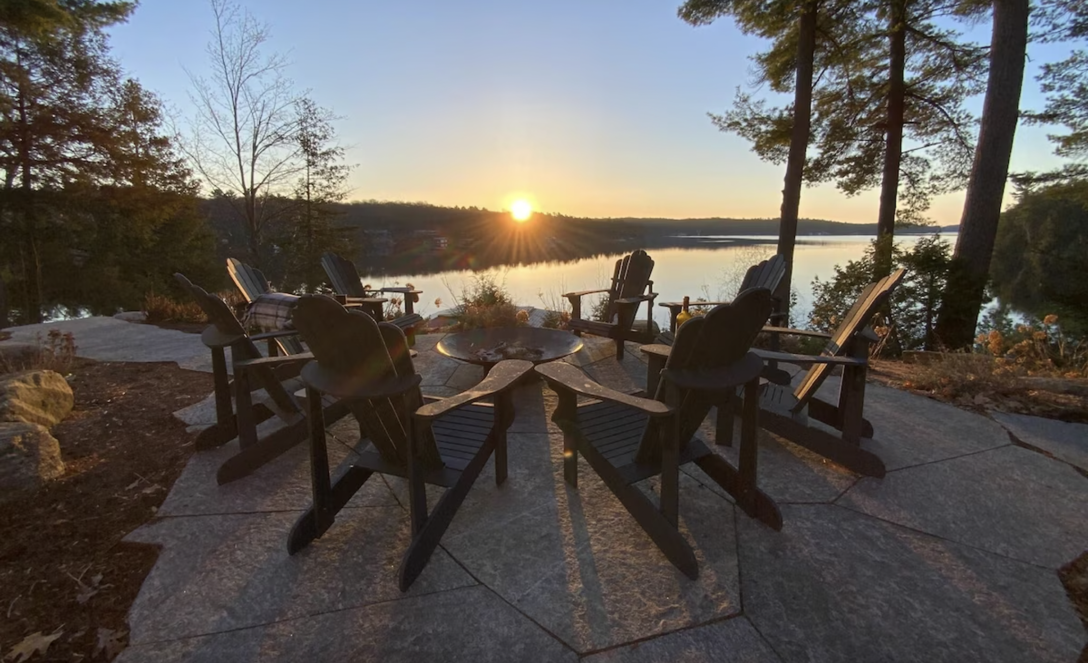 Muskoka chairs sit around a fire pit overlooking the lake at sunset